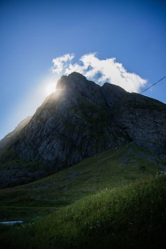 Sunlight peeks over a rugged mountain peak under a vibrant blue sky with scattered clouds, creating a serene natural landscape.