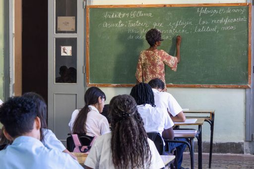 A teacher writes on a green chalkboard in a Cuban classroom setting.