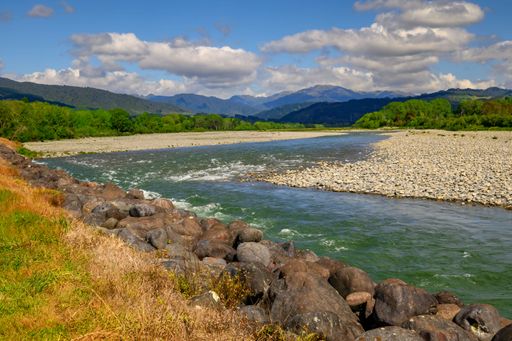 Scenic view of a river flowing through a rocky landscape with lush greenery and mountains under a cloudy sky.