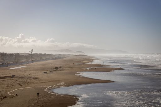 Scenic view of a vast sandy beach with gentle waves and a distant windmill under a clear sky.