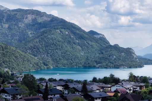 Scenic view of an alpine lake surrounded by lush green mountains and a quaint village under a partly cloudy sky.