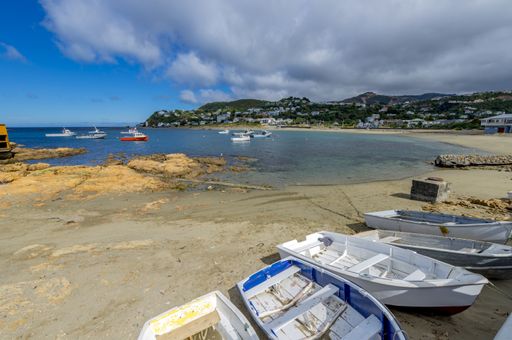 Row boats are tied up on the beach while fishing boats bob in the calm water of Island Bay in New Zealand's Wellington. Houses dot the hills behind.