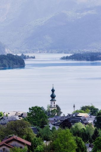 Scenic view of a charming lakeside village with a church tower, surrounded by lush greenery and mountains.