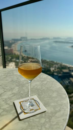 A glass of orange wine on a marble table with a cityscape and ocean view in the background, capturing a serene urban setting.