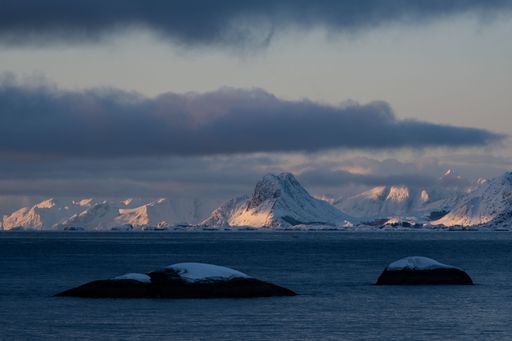 Scenic view of snow-capped mountains and ocean under a dramatic cloudy sky at sunset in Norway.