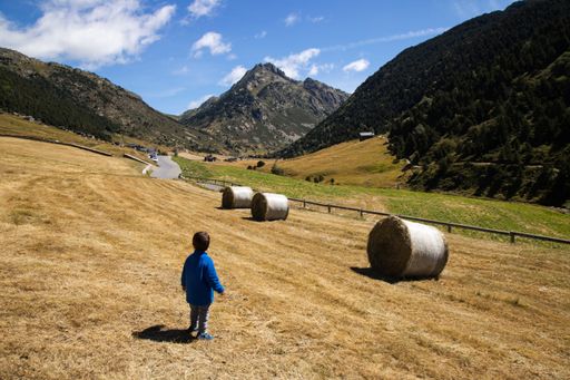 A young child in a blue jacket stands in a picturesque mountain valley with hay bales and a clear sky, capturing the beauty of nature.