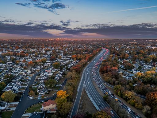 Aerial view of a suburban neighborhood with a busy highway at sunset, showcasing autumn foliage and distant city skyline.