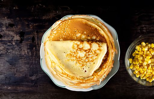 A top view of a stack of pancakes on a white plate next to a bowl of corn kernels