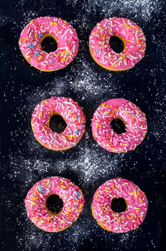 Top view of six pink frosted donuts with sprinkles on a dark surface dusted with powdered sugar.