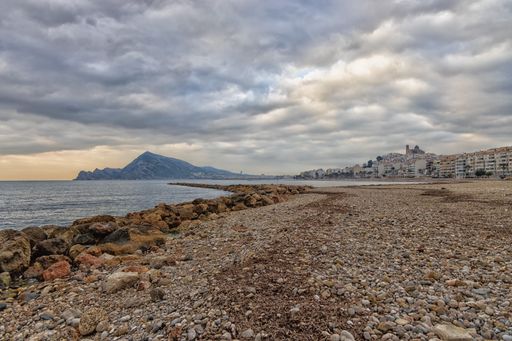 A lakeshore with stones and pebbles with a mountain and a cloudy dark sky
