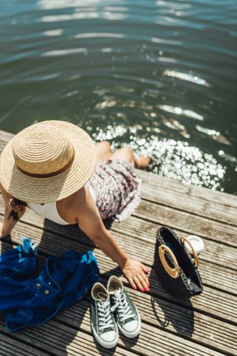 A vertical shot of a young lady sitting on a wooden dock and leaning back on hands by a lake