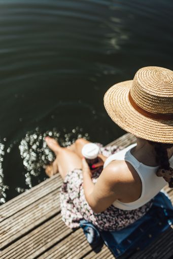A vertical shot of a young lady holding a cup of coffee and sitting on a wooden dock by a lake