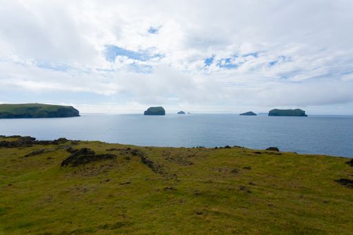 Scenic view of the Vestmannaeyjar islands from a grassy cliff in Iceland under a partly cloudy sky.