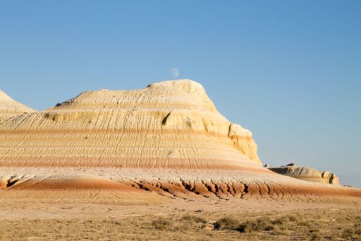 Scenic view of a striped desert hill under a clear blue sky with the moon visible, showcasing unique geological formations.