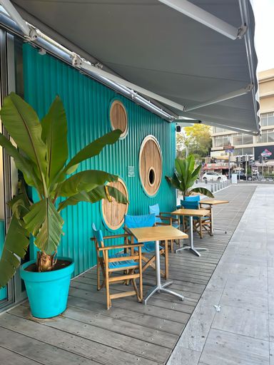 Outdoor cafe seating with wooden tables and blue chairs against a vibrant teal wall, featuring circular windows and potted plants.