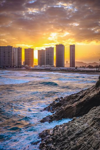 Sunset over a coastal cityscape with high-rise buildings and rocky shore, waves crashing against the rocks, dramatic clouds in the sky.