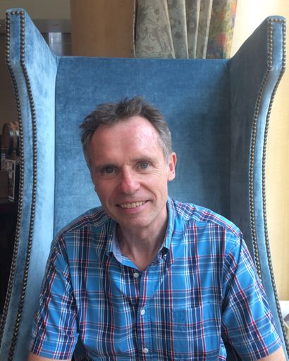 A vertical shot of a Caucasian man sitting on a big blue velvet chair and smiling into the camera