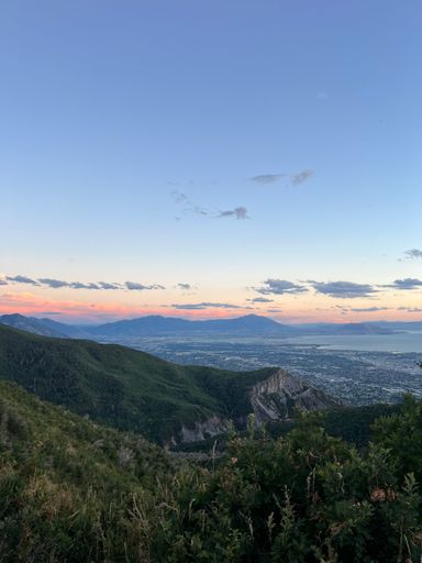 A stunning view of a lush green mountain range under a clear blue sky during sunset, overlooking a cityscape and a distant lake.