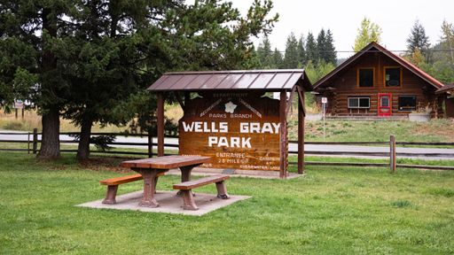 Wells Gray Park entrance sign with a wooden picnic table and a rustic cabin in the background, surrounded by lush greenery.