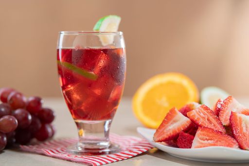 Close-up of glass cup with homemade summer wine in foreground next to plate of chopped strawberries against light brown background