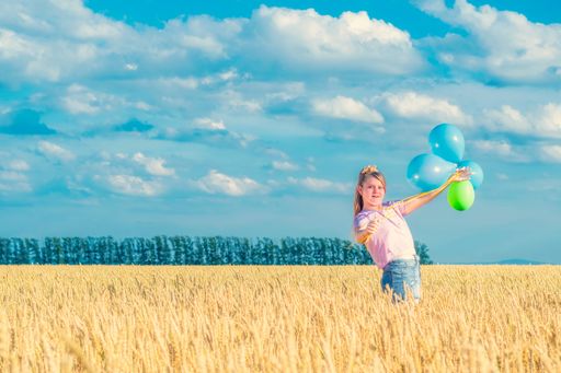 A young girl with colorful balloons in her hands is standing among a yellow wheat field against a blue cloudy sky. Looks into the camera.