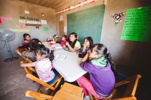 A group of kids sitting by a table and listening to their teacher in a small Mexican village church