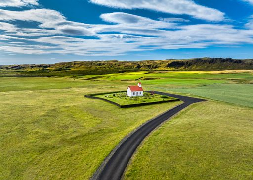 Scenic aerial view of a solitary white house with a red roof in a vast green landscape under a bright blue sky, Iceland.