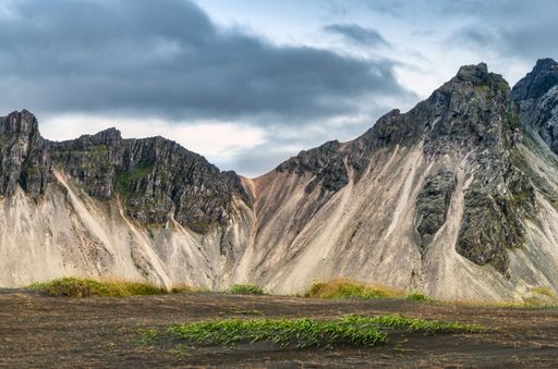 Dramatic Icelandic mountains under a cloudy sky with green patches on the black sand foreground.