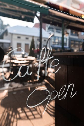 A cafe window with 'Caffe Open' written in white, reflecting an outdoor seating area in soft sunlight.