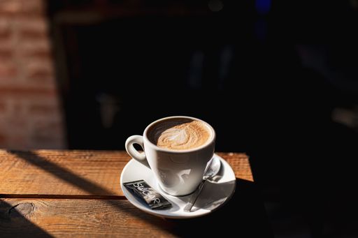 A warm cup of cappuccino with latte art sits on a wooden table in a cozy cafe setting, with sunlight casting gentle shadows.