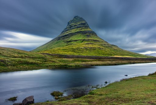 Scenic view of Kirkjufell mountain with a calm reflective lake under a dynamic cloudy sky in Iceland.