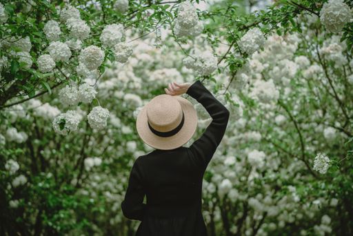 A woman in a straw hat stands amidst blooming white flowers, surrounded by lush greenery in a serene garden setting.