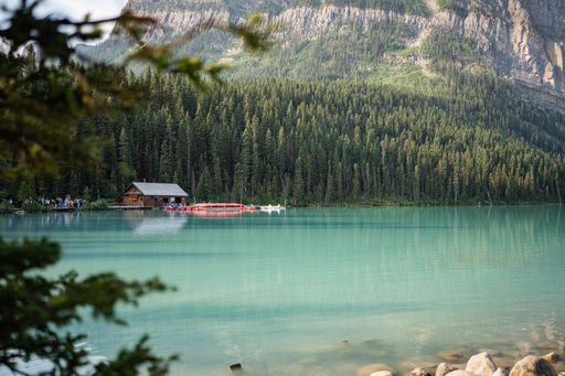 Scenic view of a serene lake with a boathouse and canoes, surrounded by lush pine forest and mountains in Banff National Park, Alberta, Canada.