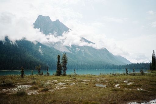 Scenic view of a serene lake with a towering mountain and misty clouds in the background, surrounded by lush greenery.