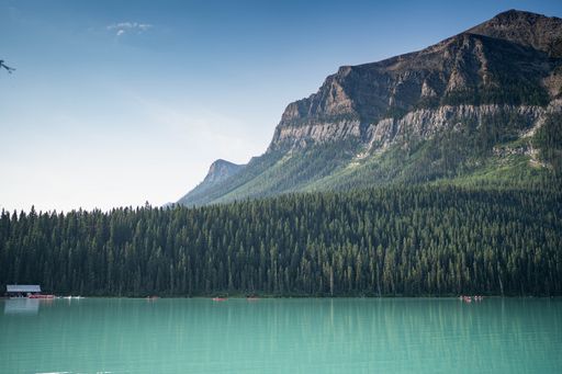 Scenic view of a serene turquoise lake surrounded by dense forest and towering mountains under a clear blue sky.