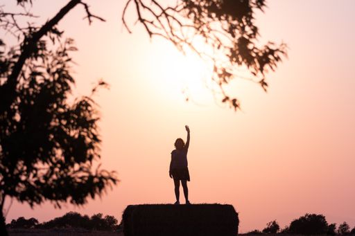 The silhouette of a woman waving her hand on top of a hay bale under a bright sky at sunset in the countryside