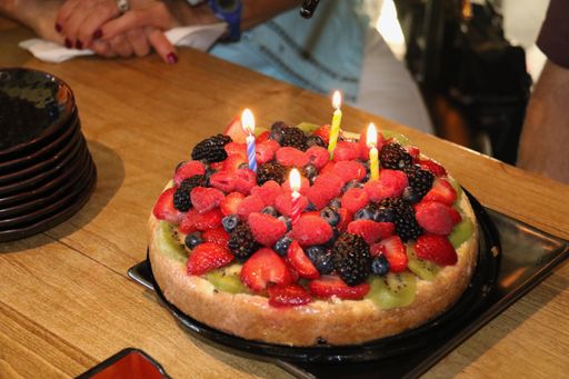 Colorful fruit birthday cake with lit candles on a wooden table, surrounded by people celebrating.