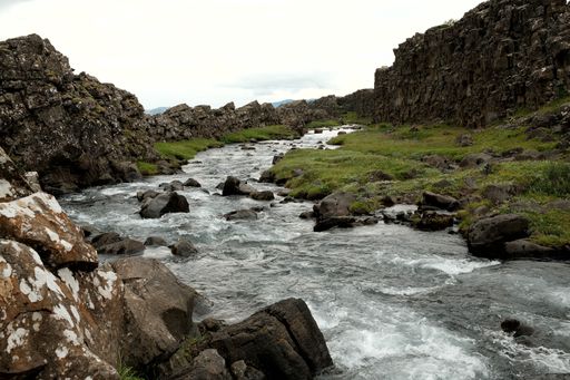 Scenic view of a rocky river flowing through a lush green valley in Thingvellir National Park, Iceland, under a cloudy sky.