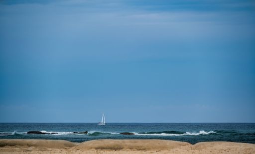 A serene ocean view with a sailboat on the horizon under a clear blue sky, captured from a sandy beach.