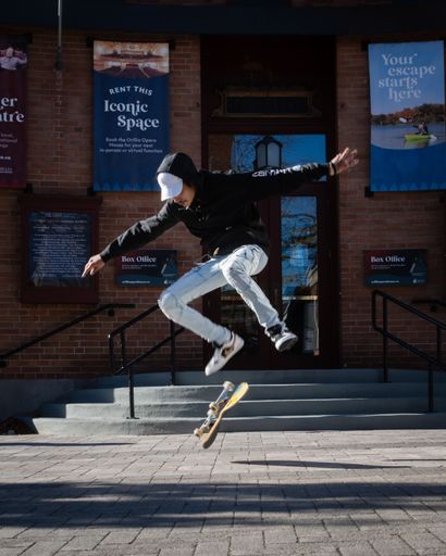 Young male skateboarder in bright sun doing flat land flip tricks on his skateboard in front of public library in Orillia