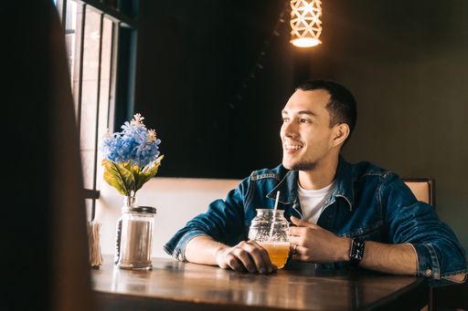 A cheerful young Hispanic man holding a refreshing drink and looking out the window in a restaurant