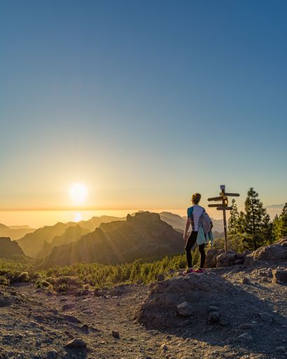 A woman in her 30s enjoys a scenic mountain view at sunset, standing near a signpost, with a clear sky and lush forest below.