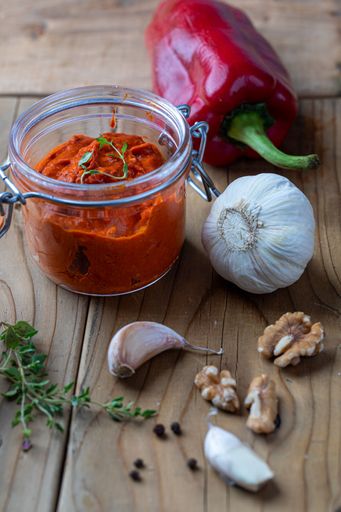 A jar of vibrant red pepper sauce with fresh ingredients like garlic, walnuts, and herbs on a rustic wooden table.