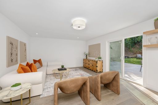 Modern living room with white walls, orange cushions, and wooden furniture, featuring a large glass door opening to a patio.