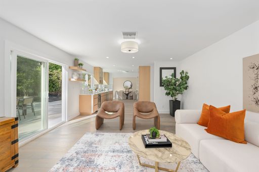 Modern open-concept living room with natural light, featuring a white sofa, orange pillows, and wooden accents, overlooking a patio.