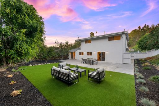 Modern backyard with artificial grass, patio furniture, and a white house under a vibrant sunset sky.