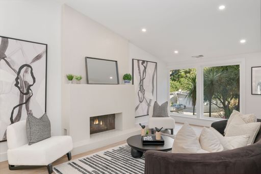 Modern living room with a minimalist design featuring a fireplace, abstract art, and large windows overlooking greenery.