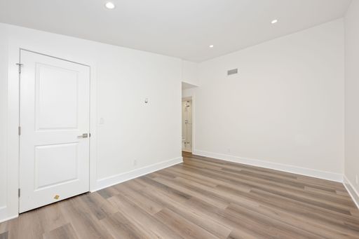 Minimalist empty room with white walls and wooden floor, featuring a closed door and a glimpse of a bathroom.