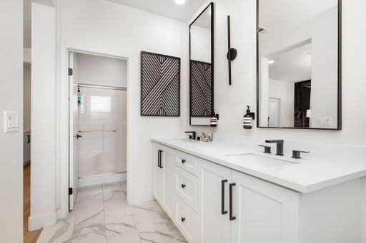 Modern bathroom interior with dual sinks, black fixtures, and geometric wall art, featuring white cabinetry and marble flooring.