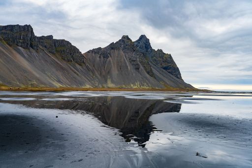 Stunning view of Vestrahorn mountain mirrored in the calm waters of a black sand beach in Iceland under a cloudy sky.